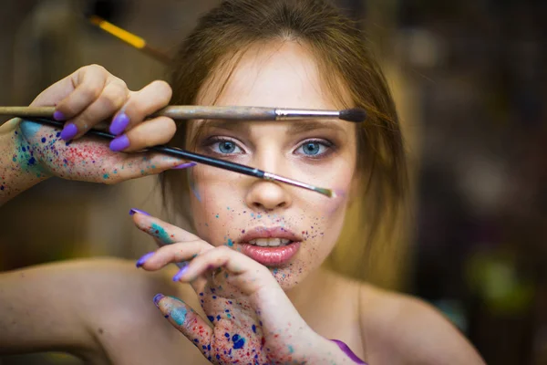 Close-up van portret van mooie vrouwelijke artiest met vuile handen met verschillende verven op hen, verf penselen in de buurt van haar gezicht en ogen houden. — Stockfoto