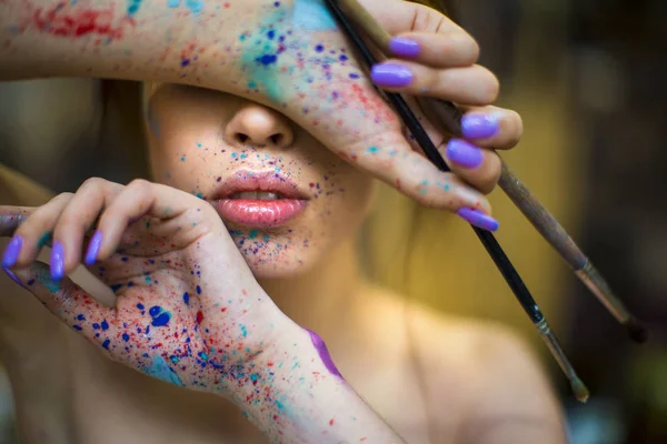 Close up portrait of beautiful female artist with dirty hands with different paints on them, holding paint brushes near her face and eyes. — Stock Photo, Image
