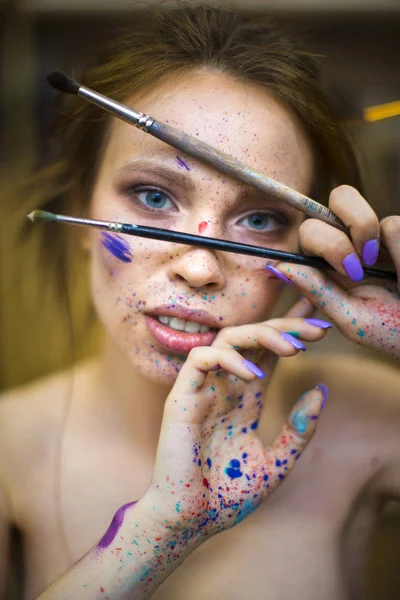 Ritratto ravvicinato di bella artista femminile con mani sporche con diverse vernici su di loro, tenendo pennelli vicino al viso e agli occhi . — Foto Stock