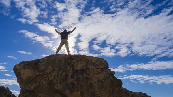 Happy sporty man with backpack and raised up arms standing and on the rock and looking at the seashore and mountains at sunset in summer. Man, sea, mountain ridges and orange sky — Stock Photo, Image