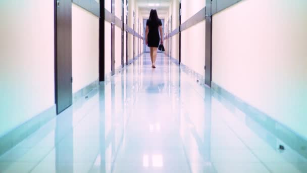 Vista trasera de una mujer en un vestido negro caminando por el pasillo del hotel. Ella sostiene los zapatos en su mano — Vídeos de Stock