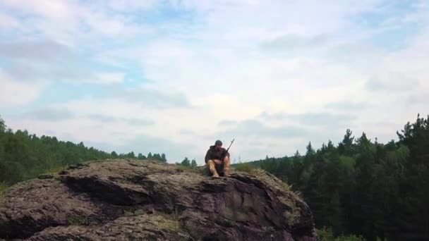 Un francotirador con un rifle se sienta en la cima de la montaña e inspecciona el territorio. Vista desde el dron. la cámara se mueve desde el soldado, revelando una vista del paisaje — Vídeos de Stock