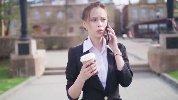 Jonge succesvolle zakenvrouw met behulp van slimme telefoon in het stadscentrum van de, professionele vrouwelijke werkgever in gesprek met zakenpartner koffie drinken. Zakelijke district wolkenkrabbers in achtergrond — Stockvideo