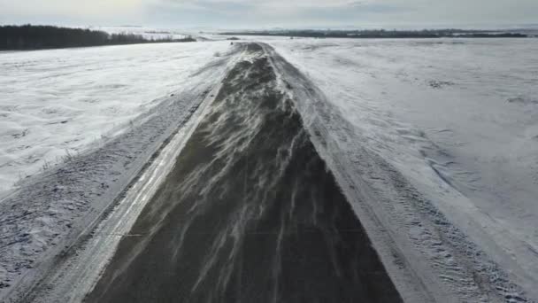 Antena: avanzando, vista desde arriba en una carretera congelada. El viento lleva nieve a través de la carretera — Vídeos de Stock