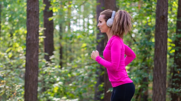 Mujer corriendo en una zona forestal y escuchando la música a través de teléfonos móviles. Vista lateral . — Foto de Stock