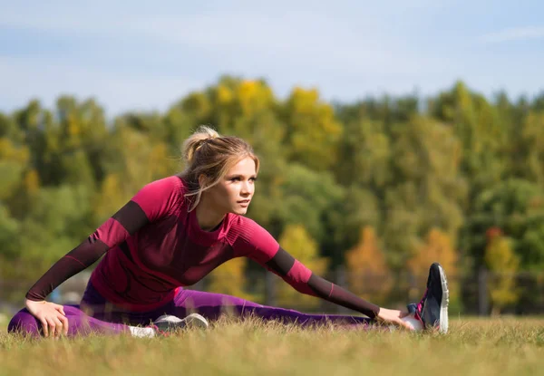 Mujer fitness 20s haciendo ejercicio y estirando las piernas mientras está sentado en un césped en el parque . — Foto de Stock