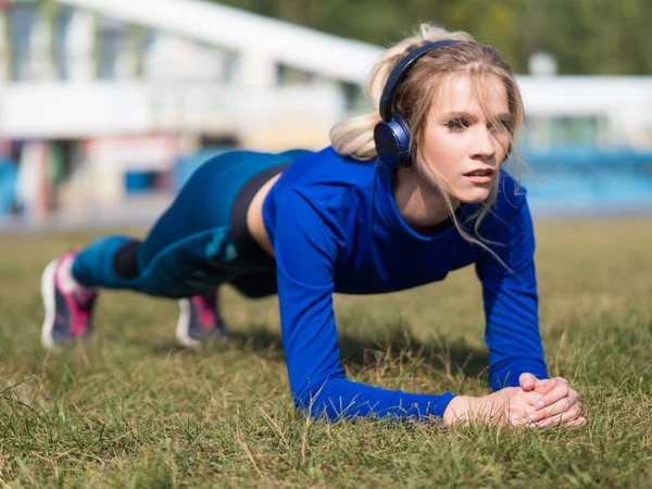 Chica en forma de auriculares haciendo ejercicio de tablón al aire libre en el parque cálido día de verano . — Foto de Stock