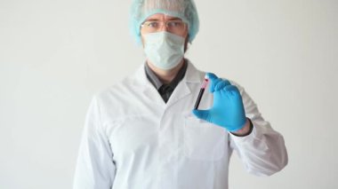 Beyaz önlüklü ve koruyucu maskeli bir doktor test tüpünde kan örneği gösteriyor ve başparmağını yukarıda tutuyor..