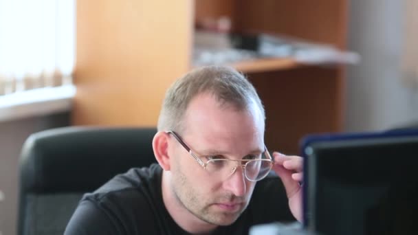 Een man werkt in een kantoor op een computer. Zijn ogen zijn moe en hij doet zijn bril af.. — Stockvideo