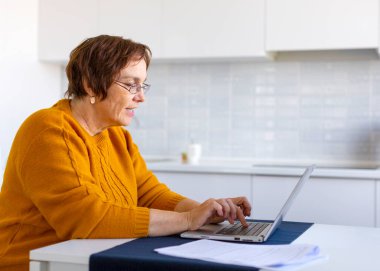 Evde dizüstü bilgisayarla çalışan gülen yaşlı kadın..