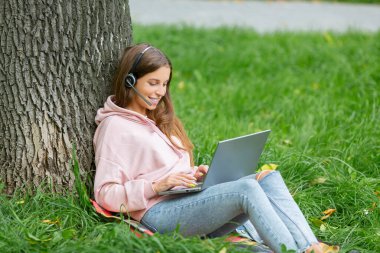 Kulaklığı ve dizüstü bilgisayarı olan gülümseyen kadın halka açık parkta çimlerin üzerinde oturuyor..