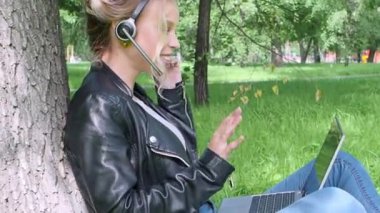 Dizüstü bilgisayarı ve kulaklığı olan gülümseyen kadın şehir parkında otururken internetten konuşuyor. Yan görünüm.