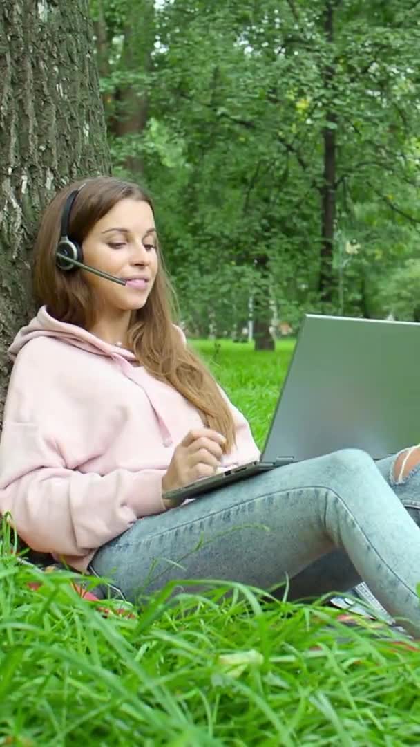Egy fiatal nő laptoppal és fejhallgatóval online beszélgetett, miközben a City Parkban ült. Függőleges videó. — Stock videók