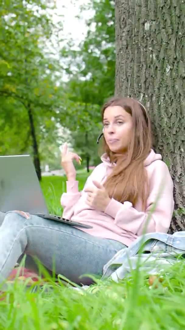 Vertikales Video - junge Frau mit Laptop und Headset unterhält sich im Stadtpark online. — Stockvideo