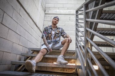 young caucasian tattooed singer rap posing sitting on the steps of a metal ladder clipart