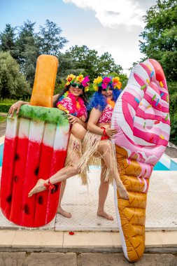 Havuzun kenarında Hawaii kostümü giymiş iki gülümseyen bayan arkadaşın komik portresi.