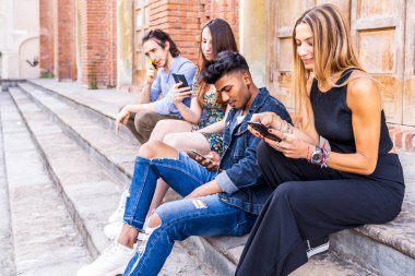 Mermer bir merdivende oturan bir grup çok ırklı arkadaş sosyal ağlardaki içeriği paylaşmak için akıllı telefonları kullanıyorlar.