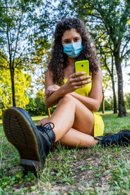 Güzel genç bir kadın, acil durum sırasında bulaşıcı olmayan bir maske takarak akıllı telefon kullanıyor.