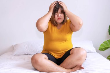 Şişman genç bir kadın, beyaz yatakta oturmuş, evde el ele tutuşuyor. Yatak odasında fazladan ağırlık çeken üzgün bir kadın. Obezite sağlıksız bir kavram.