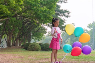 Neşeli çocuklar şirin kız çok renkli balonlar parktaki yeşil çayırda büyük ağaçlı. Neşeli çocuk, siyah saçlı, pembe elbiseli, renkli balonları olan bir kız..