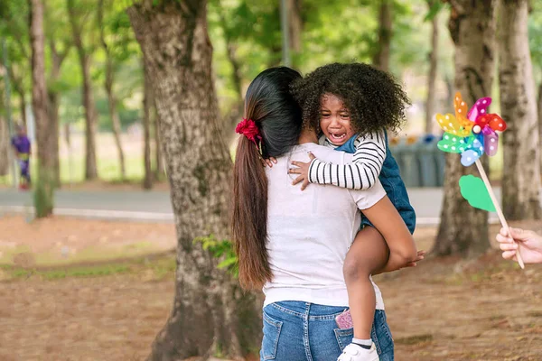 Anne parkta kızını ağlarken kucakladı. Küçük siyah kız ağlıyor annesi kollarında büyük bir ağacın altında dururken.