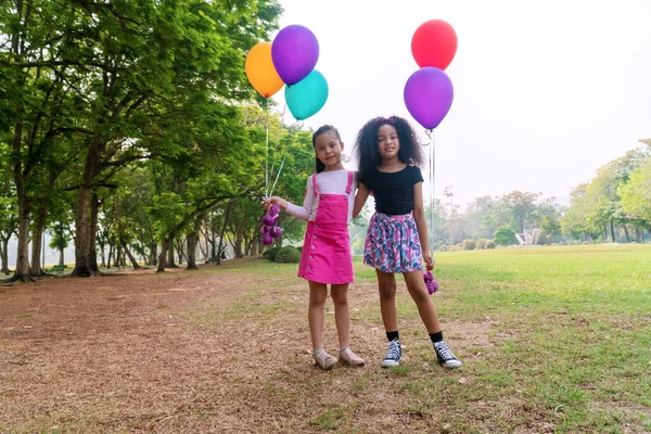 陽気2人の子供の女の子は緑の芝生の上に一緒に立っている間カラフルな風船を保持します 公園でカメラを見ながら自由を感じながら風船でかわいい女の子の遊び 友情の概念 — ストック写真
