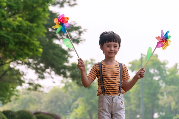 公園で朝木と一緒に立っている間 幸せな男の子は風車を手に持っています ガーデンパークで風を見つけながら松輪遊びの子供かわいい男の子 — ストック写真