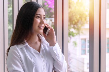 İş kadını pencerelerin yanında dururken akıllı telefondan projeden bahsediyor. Dışarıdaki bir ofise bakarken elinde telefonla gülümseyen genç bir kadın. Yaşam tarzı teknoloji konsepti.