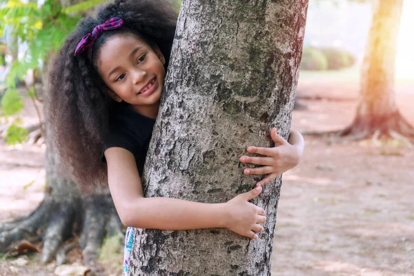 公園の木の後ろにはレースガールの巻き毛が立っていた 小さなかわいい子供は木の後ろから外を見て 公園で隠れて遊んでいます 小さな女の子手を含む大きな木上の自然 — ストック写真