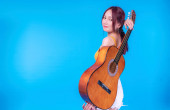 Šťastná mladá žena s akustickou kytarou za zády na modrém pozadí. Portrét veselá žena drží kytaru na zádech, zatímco stojí na izolovaném pozadí.
