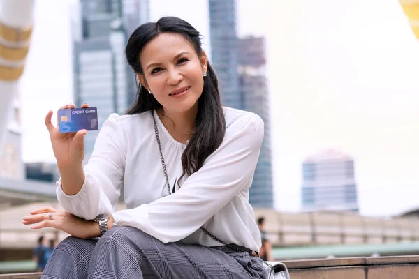 Shopper senior woman hold mock up credit card in her hands while looking at camera. Portrait customer female sitting in street city with card feeling freedom paying shopping. Lifestyle payment concept