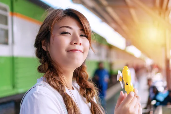 Happiness young woman sweating feeling uncomfortable from heat stroke in hot summer weather hold battery-powered fan at train station. Girl brown long hair using portable fan while standing at outdoor