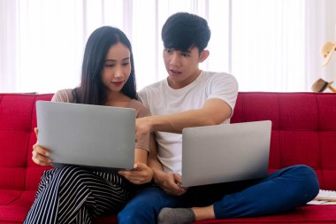 Genç çiftler yeni normal politikaların uygulanması için kırmızı koltukta otururken dizüstü bilgisayar kullanıyorlar. Salgın konseptinde teknoloji iletişimi.