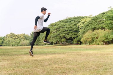 Enerji egzersizi eğitimi almış yakışıklı genç bir atlet ve halka açık parkta büyük yeşil bir ağaçla dışarı atlıyor. Aktif sağlık ve motivasyon yaşam tarzı ve hırs kavramı