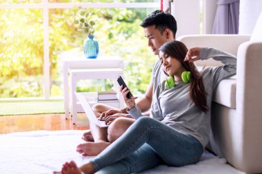 Genç bir adam elinde akıllı telefonuyla kanepede otururken internette dizüstü bilgisayar kullanıyor. Kulaklıklı genç bir kadın evde telefonla alışveriş yapıyor.. 