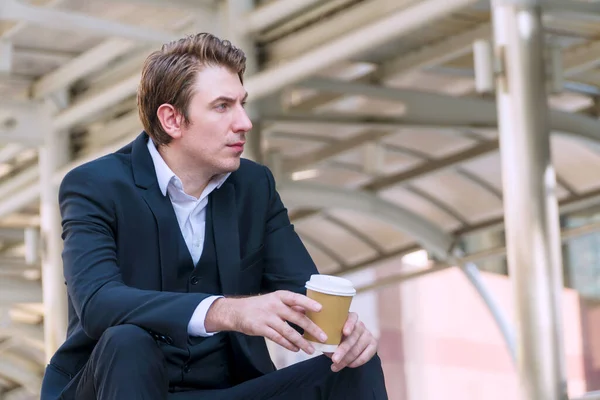 Executive handsome man wear suit holding cup of coffee while sitting  alone in morning at urban city. Businessman looking at something while thinking about work on street. Business concept.