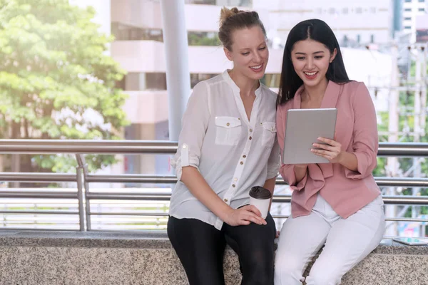 Two young businesswoman holding tablet in hand while looking at something together. Smile young girl holding tablet with her colleague hold cup of coffee while sitting in city. Communication concept.