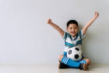 Neşeli küçük çocuk yerde otururken futbol forması giyer. Futbol futbolu oynayan mutlu okul çocuğu, gülümseyerek oyununu oynuyor. Sağlıklı konsept.