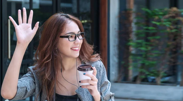 Attractive young woman sitting outdoor in cafe while waving hand saying hello her friend. Business woman wear glasses holding cup of coffee while waving hands greeting her friend say hi or goodbye.