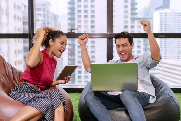 Happy young businesspeople colleagues sitting on sofa with notebook while raise fist up together felling happy their planning job successful in office on city background. Teamwork concept.