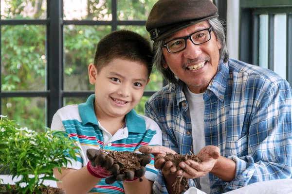 活動内容かわいい男の子とおじいさんが緑の家で土を手にしながら孫に木を育てる笑顔のおじいさんが家で土を手にしながら教えてくれる — ストック写真
