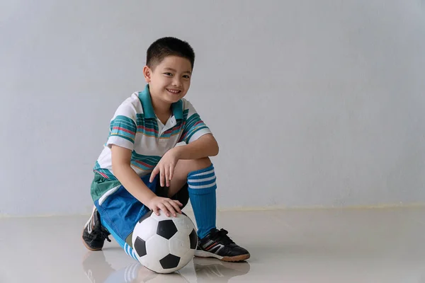 カメラを見ながら床に座ってサッカーと陽気なかわいい男の子 女子高生は部屋に座っている間 ボールの笑顔でサッカーのスポーツウェアを着ています ライフスタイルキッズウェルネスコンセプト — ストック写真
