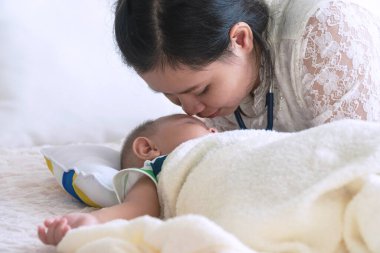Mutlu genç anne, oğlu yatakta uyurken onun tatlı bebek yüzünü sevgiyle öpüyor. Anneler günü konsepti.