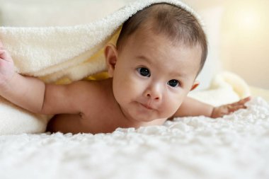 Neşeli şirin bebek beyaz battaniyenin altında kameraya bakıyor. Masum bebek, beyaz yatakta kafasında havluyla sürünüyor..