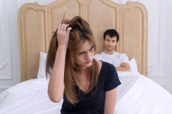 Unhappy young couple having a problem in the bedroom. Upset young wife sitting on the edge of the bed headache, against her husband lying on the bed at home. Relationship problem concept.