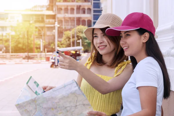 旅行者の2人の若い女性の手をつないで地図を保持しながら 方向を指す 陽気な若い女の子は手に地図を保持しながら 彼女の友人を探しているために通りに指ポイントを使用帽子を着用 休日のコンセプト — ストック写真