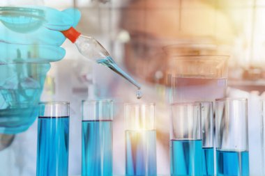 Araştırmacılar mavi kimyasalla doldurulmuş test tüplerinin üzerinde mavi bir sıvı bulunan laboratuvar pipeti tutuyorlar. Laboratuvarda koruyucu gözlüklerle çalışan genç bir bilim kadını. Çift pozlama biçimi