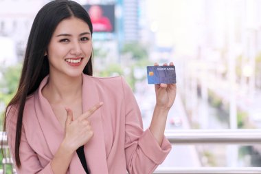 Elinde kredi kartı olan gülümseyen genç bir kadın şehrin arka planındaki kameraya bakıyor. Çekici bir iş kadını ödeme özgürlüğüyle tatmin olur ve mutlu olur..