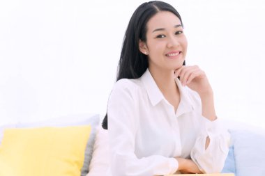 Yaşam tarzı rahatlama konsepti. Neşeli, gülümseyen genç bir kadın beyaz koltukta oturuyor. Yastık ve sarı kitapla evde oturuyor.