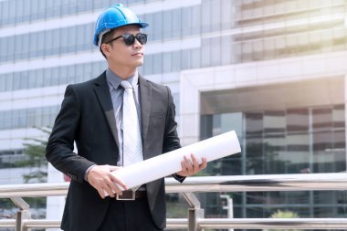 İnşaat konsepti. Başarı mühendisi güvenlik için mavi kask takar ve bina planlarını kontrol eder. Genç işadamı güneş gözlüğü takıyor. Şantiyede çalışıyor..